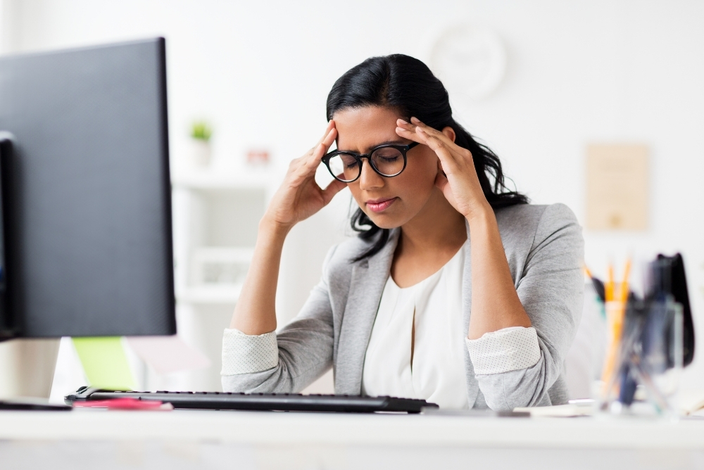 A woman with hands on the forehead and looking down on the computer