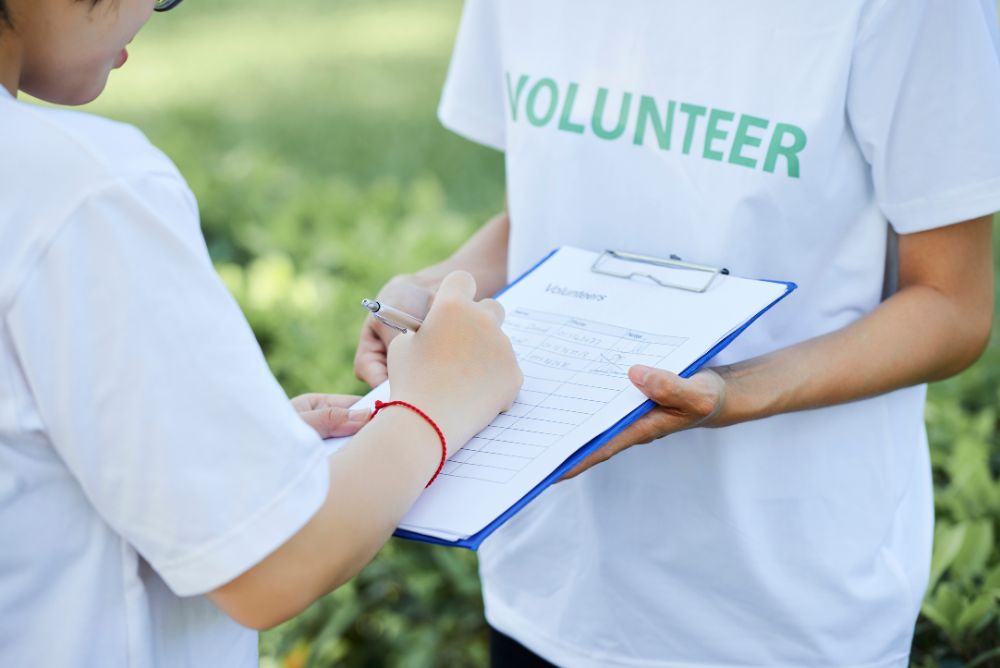 a person wearing a tshirt which says Volunteer and holding a writing board in his hand to sign up volunteers