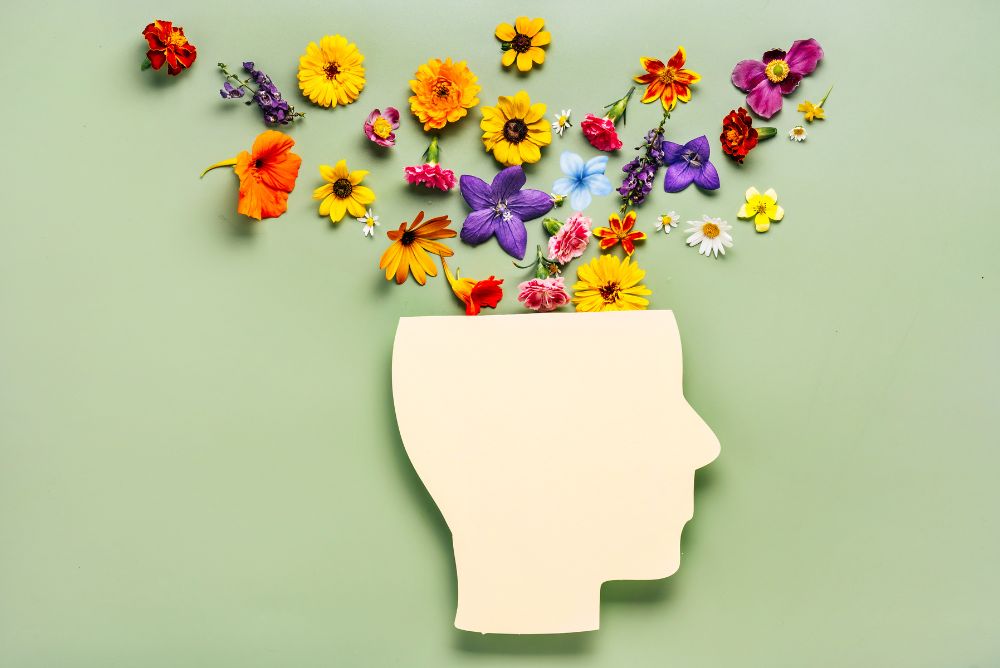 Silhouette of a human head in profile made from yellow paper. There is an assortment of colorful flowers arranged above the head to look like they are blossoming from the mind